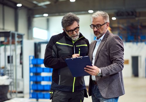 Two Professionals Reviewing a Clipboard in a Modern Industrial Warehouse Setting A manager and employee discussing details in an industrial workplace near storage units.