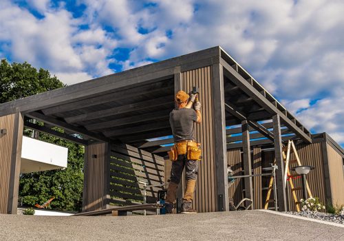 Craftsman Constructs a Modern Outdoor Structure on a Sunny Day Amid Fluffy Clouds in a Residential Area A skilled worker is building an outdoor structure, focusing on detailed wooden framing under a bright sky with clouds. The area showcases modern residential design.