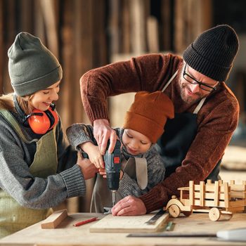Happy family doing woodwork together in workshop Positive mom and dad in aprons helping little son using drill while making wooden toys in carpentry workshop together