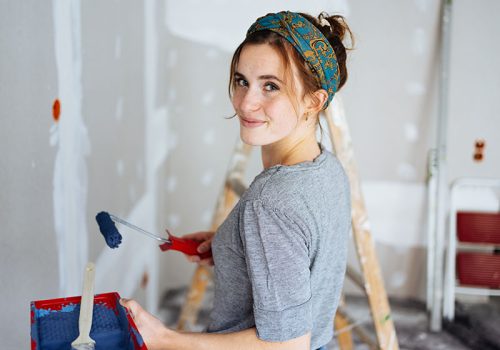 Happy young woman decorating the walls of her apartment Happy young woman decorating the walls of her apartment turning to look over her shoulder at the camera with a friendly smile