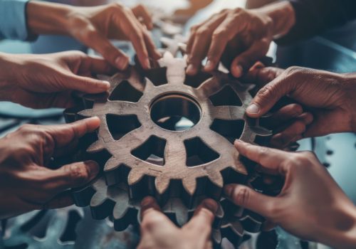 A group of hands hold a set of interlocking gears, symbolizing the concept of teamwork and collaboration. The image represents the importance of each individual contributing their part to achieve a common goal.